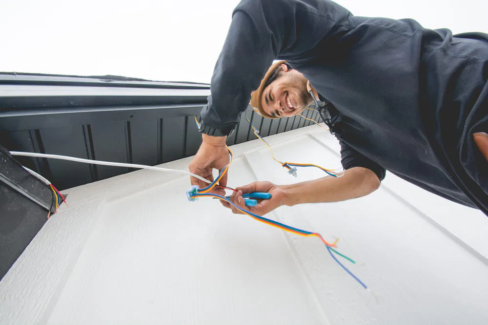 A person smiling while installing or repairing electrical wiring on an exterior wall, holding colorful wires and using tools, viewed from a low angle.