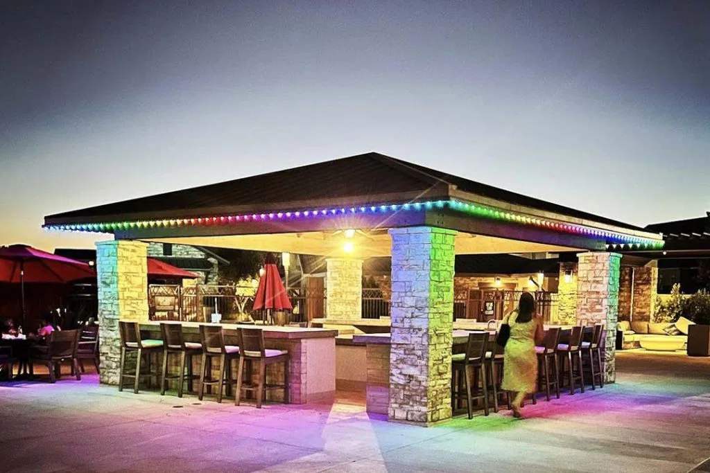 An outdoor bar with stone pillars and a dark roof is lit by multicolored LED lights at dusk. Wooden barstools surround the bar, and a person walks nearby. Red umbrellas and seating are visible in the background.