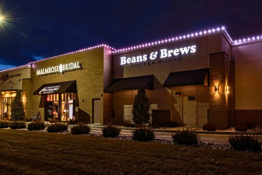 A well-lit storefront at night features two businesses: Malmrose Bridal, with a black awning, and Beans & Brews Coffeehouse, both with illuminated signs. Low shrubs and grass line the walkway in front of the buildings.