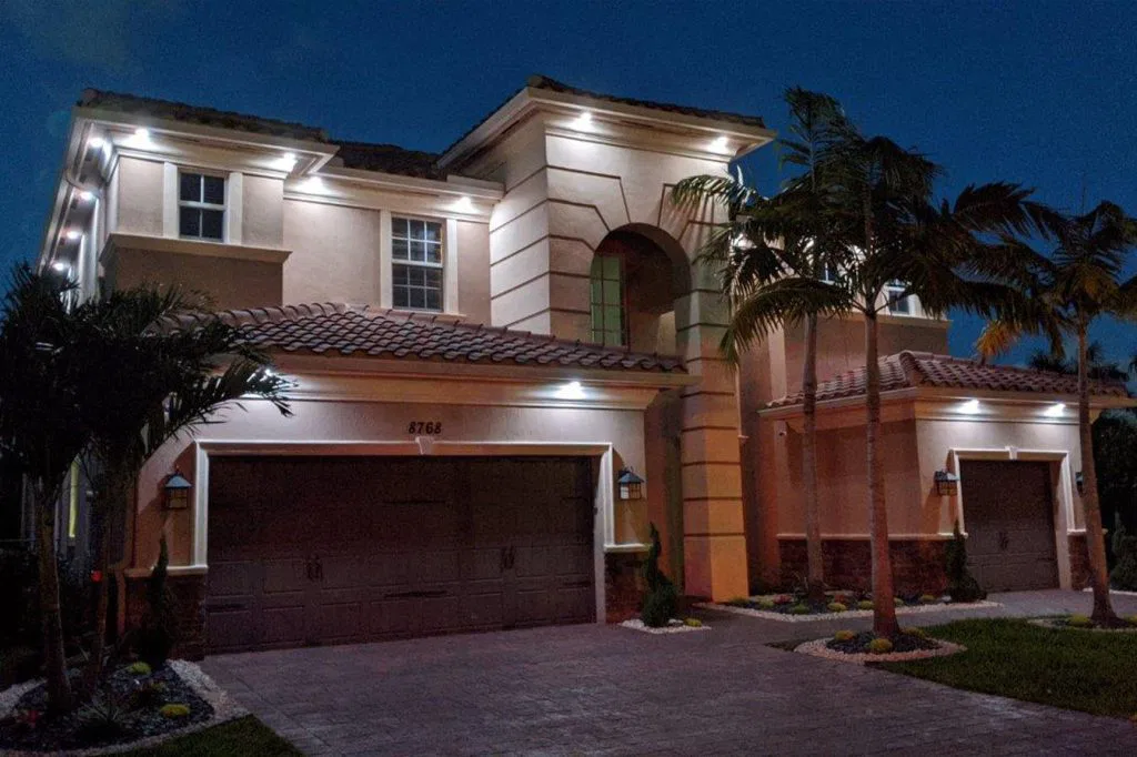 Attached image file A modern two-story house with exterior lights illuminating the beige walls and brown tiled roof at night, surrounded by palm trees and neatly landscaped greenery.