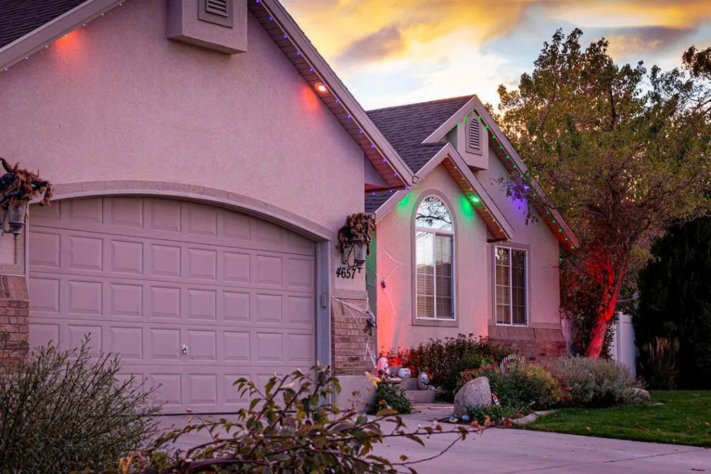 Attached image file Single-story house with a two-car garage, front yard landscaping, and colorful holiday lights along the roofline at sunset. Trees and bushes are visible near the driveway and house entrance.