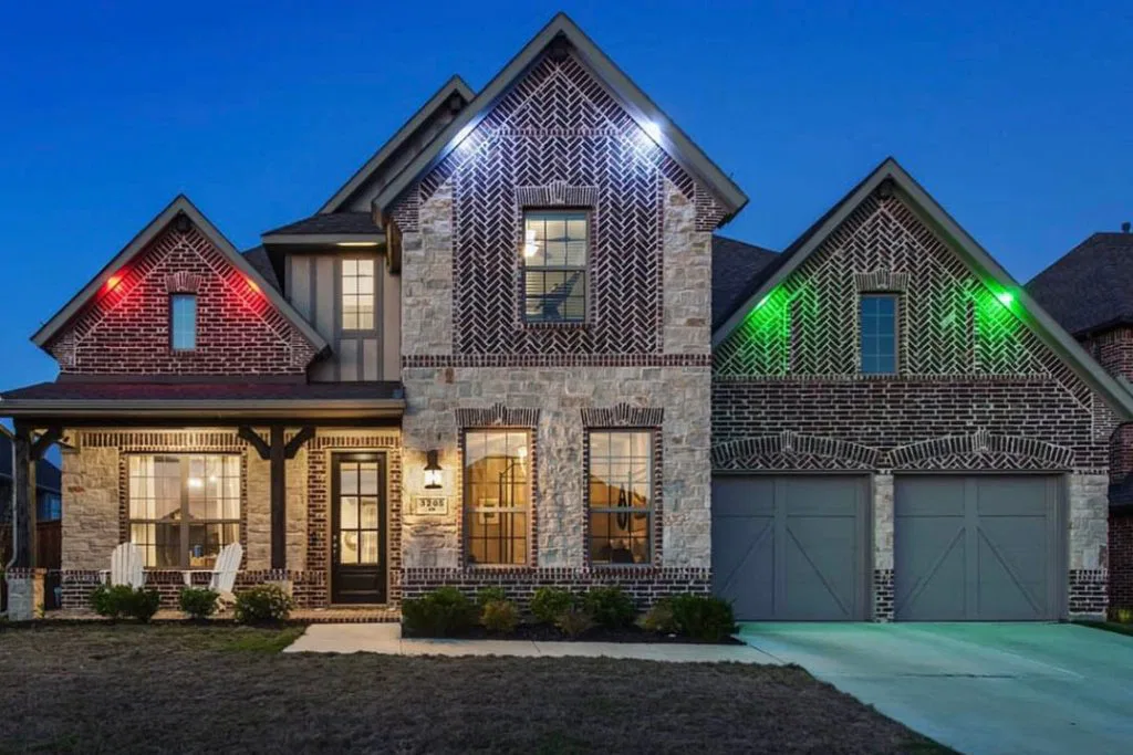 Attached image file A two-story brick and stone house at dusk features a covered porch with white chairs, double garage, and glowing red, blue, and green exterior lights, with windows warmly lit from inside.