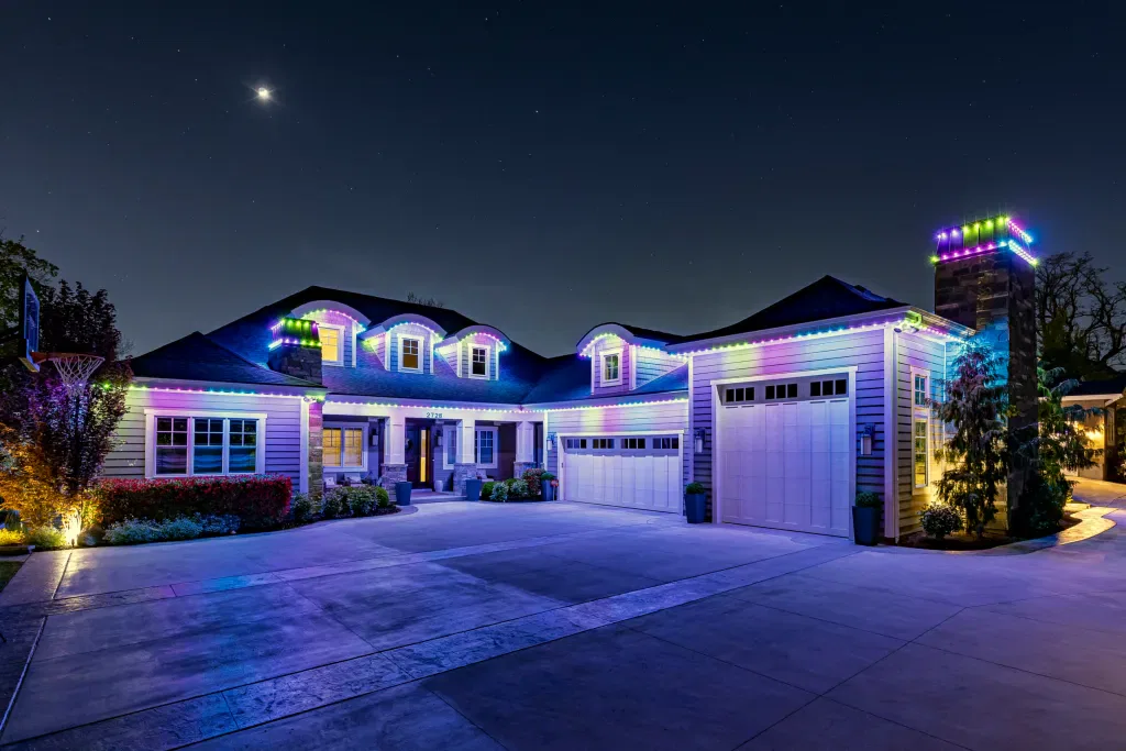 A large house at night is decorated with colorful LED lights outlining the roof, garage, and chimney. The driveway is empty and a crescent moon shines in the sky above.