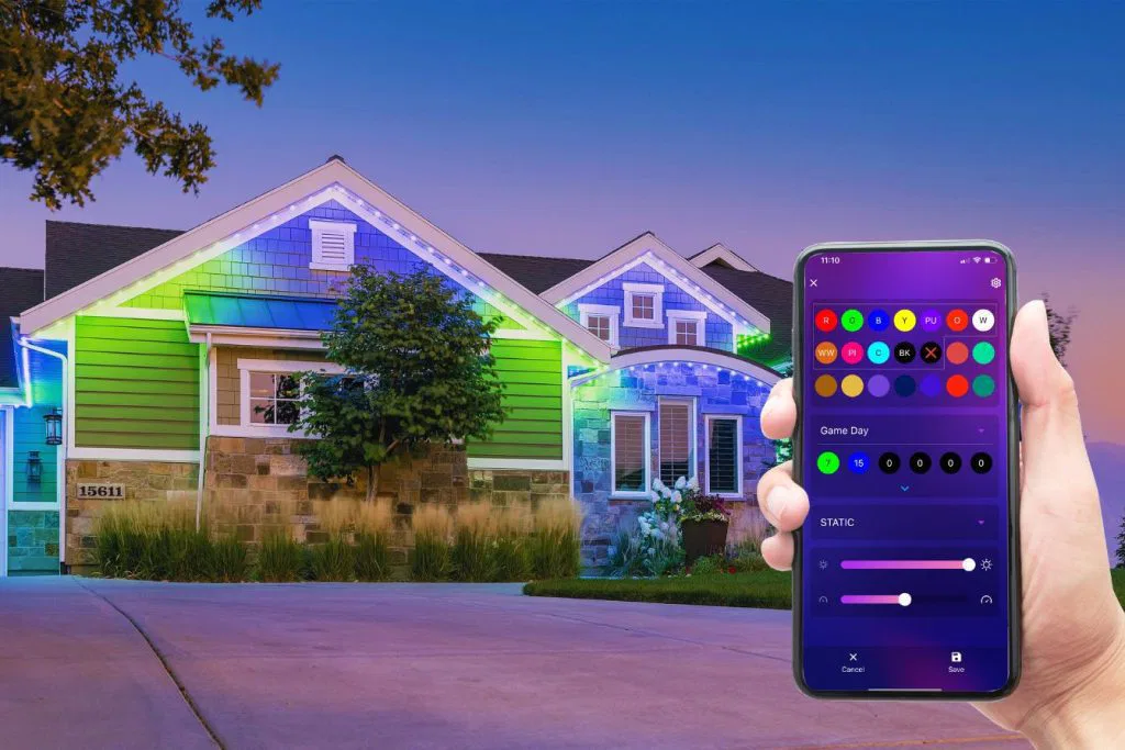 A hand holds a smartphone displaying an app for controlling outdoor house lights, which are illuminated in colorful patterns on the exterior of a modern suburban home at dusk.