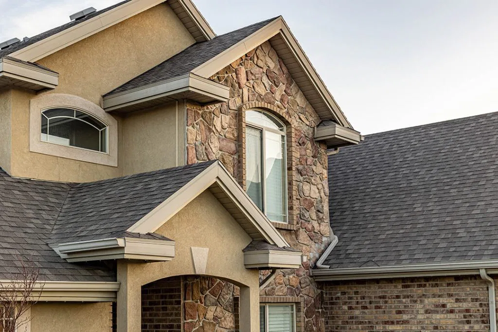 Modern two-story house exterior featuring a mix of stone and stucco siding, large windows, gable roofs with grey shingles, and partial view of a brick wall.