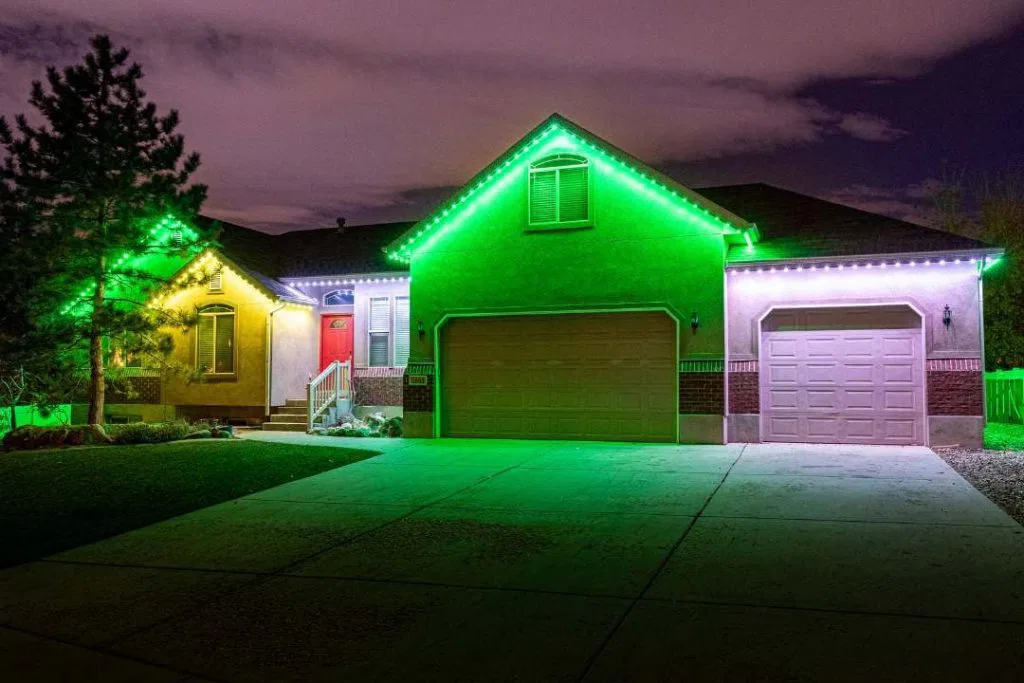 A single-story house at night is lit with green and white LED lights along the roofline, highlighting the garage doors, entryway, and front yard. The sky is cloudy, and a tree stands on the left side of the house.