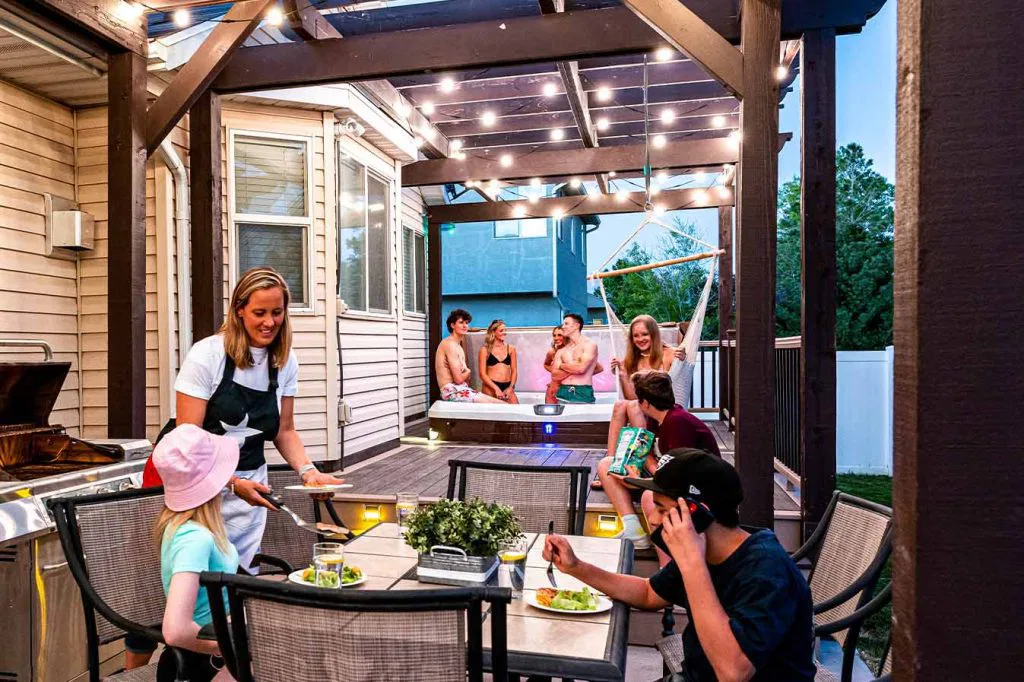 A woman serves food at an outdoor patio table to two children, while four adults relax and chat in a hot tub nearby. String lights hang overhead, and the scene is cheerful and lively in a backyard setting.