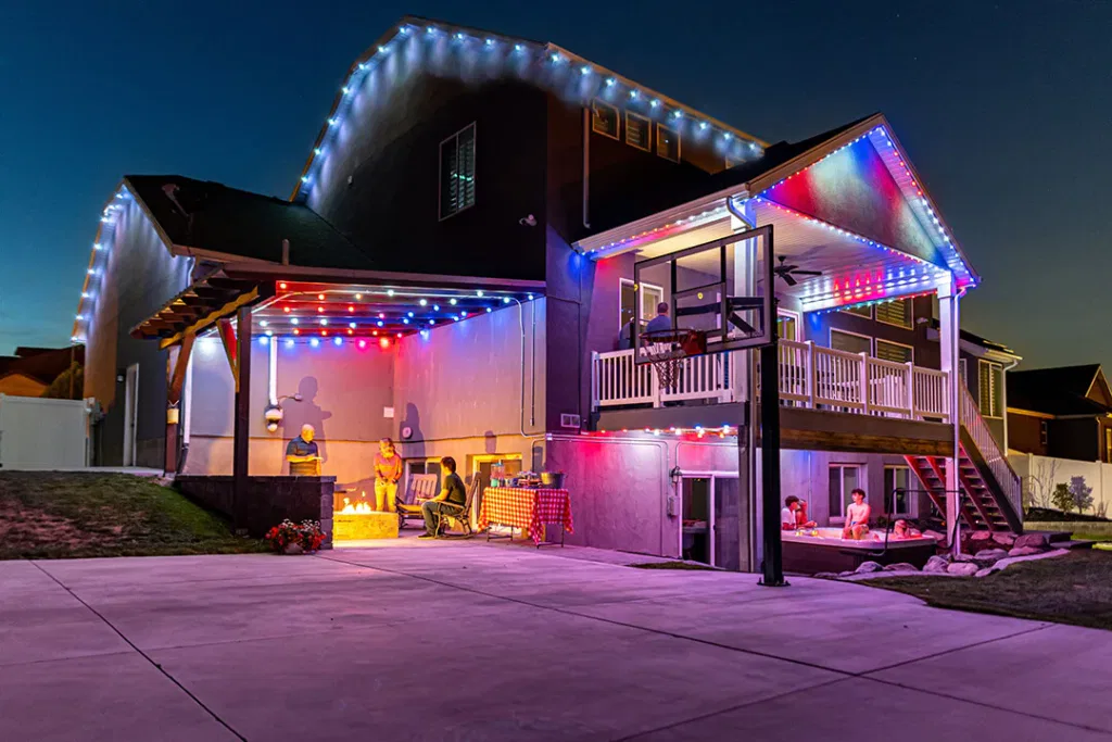 A two-story house at dusk decorated with colorful string lights. People are gathered on the patio and under the balcony, enjoying an outdoor social event next to a basketball hoop.