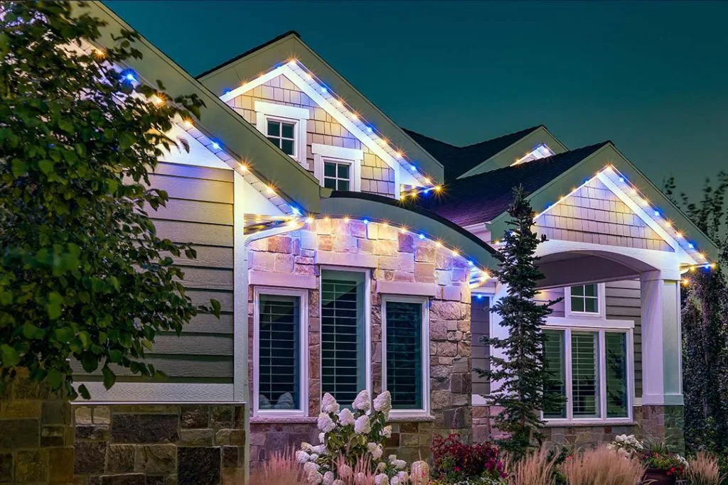A modern house at dusk with a stone and siding exterior, white-trimmed windows, landscaped garden, and blue and white string lights outlining the roof.