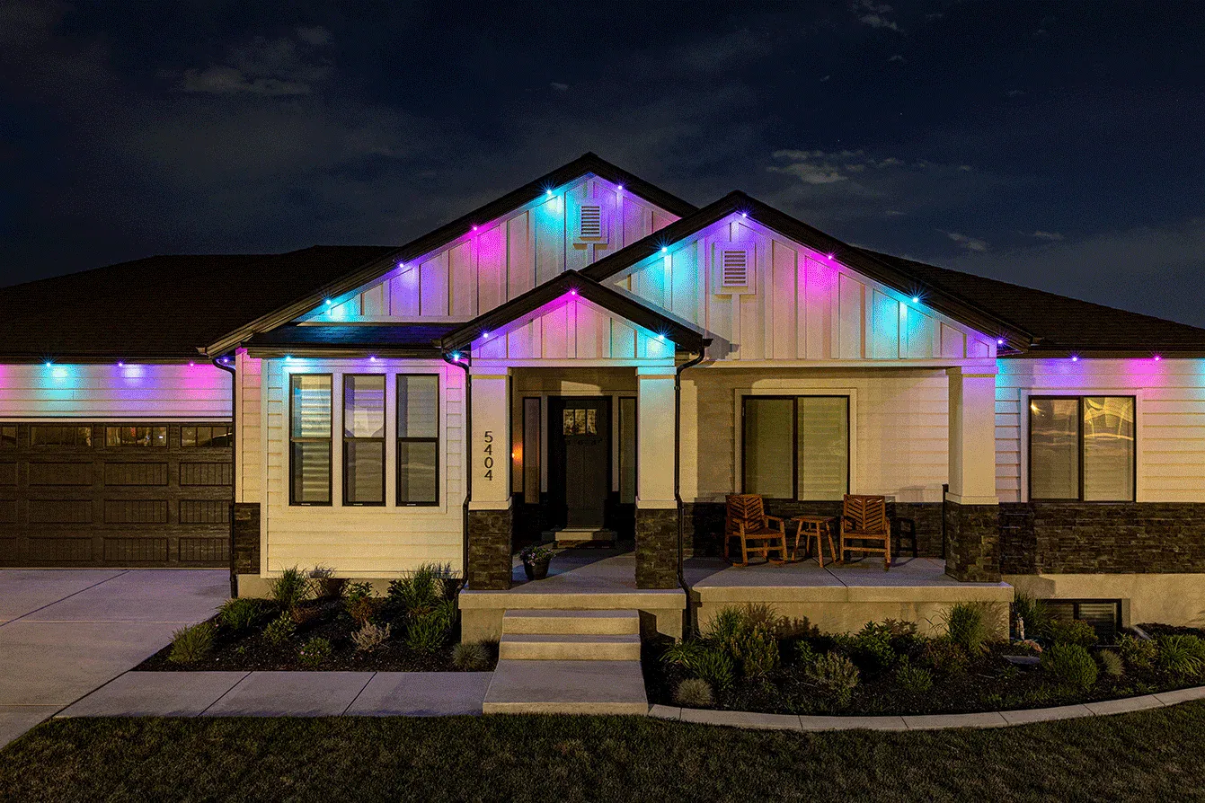 A modern single-story house at night, decorated with colorful blue, pink, and purple lights along the roofline. The front porch has two wooden chairs and is surrounded by neatly trimmed bushes.