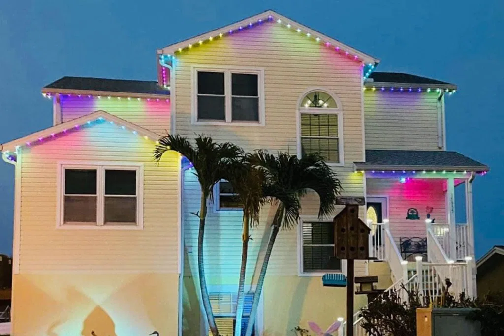 A two-story house is decorated with colorful string lights along the roof edges, glowing in the evening. Two palm trees stand in front, and a birdhouse is visible near the staircase on the right.