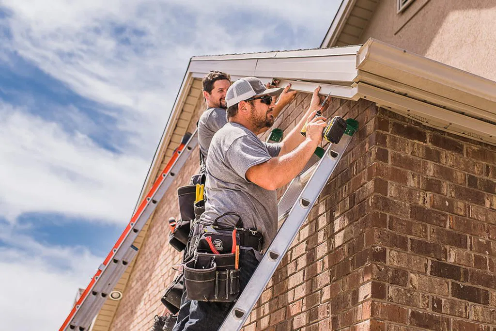 Two workers wearing tool belts stand on ladders, installing or repairing gutters on the roof edge of a brick house under a partly cloudy sky.