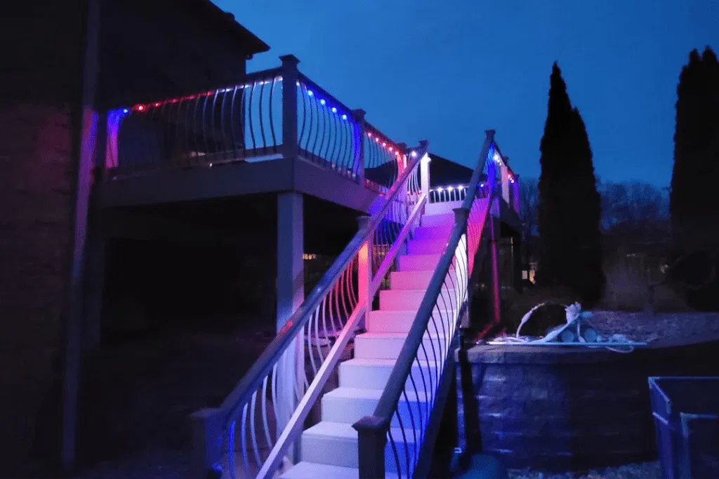 A staircase leading to an elevated deck is illuminated by red, white, and blue lights at night, creating a vibrant glow on the railings and steps. Trees and a garden area are visible in the background.