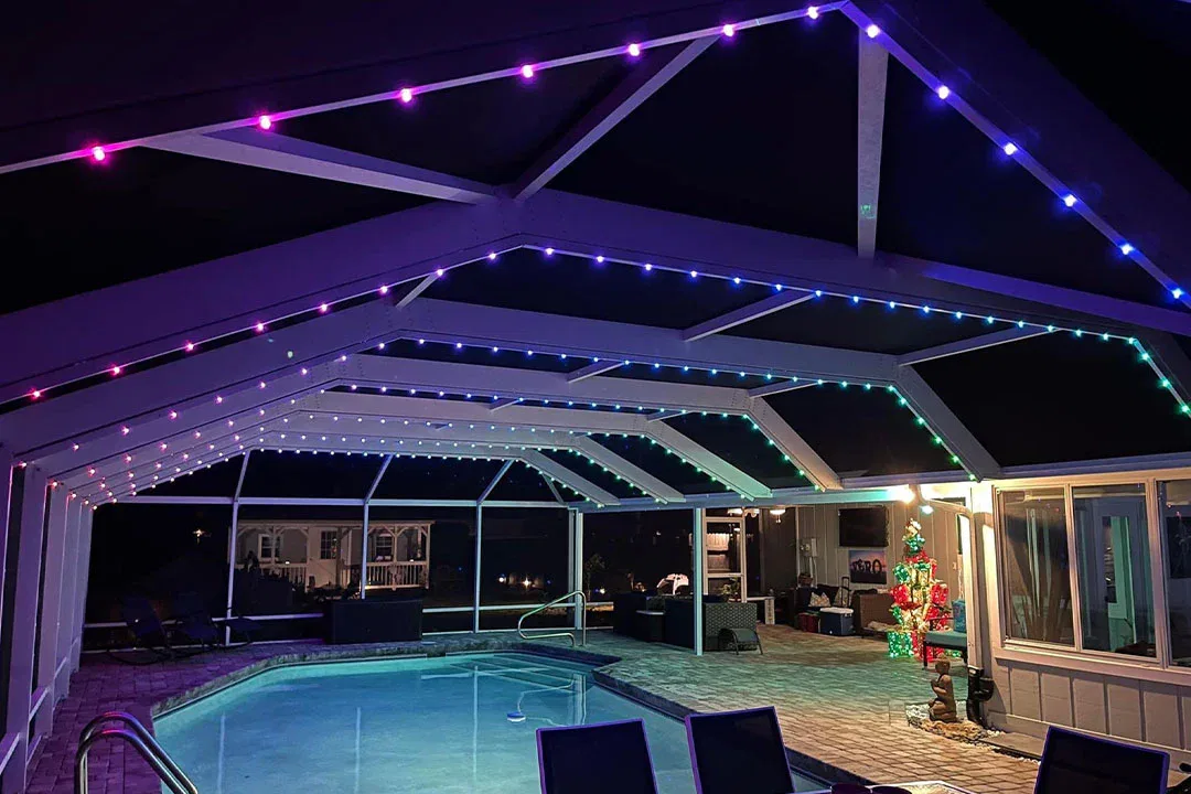 A covered outdoor pool area at night is decorated with colorful LED strip lights along the roof. There is patio furniture around the pool and a lit Christmas tree near the back.