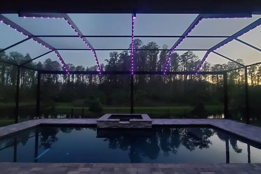 A screened-in pool area at dusk, featuring purple string lights outlining the frame, with trees and their reflection visible in the water and a sunset sky in the background.