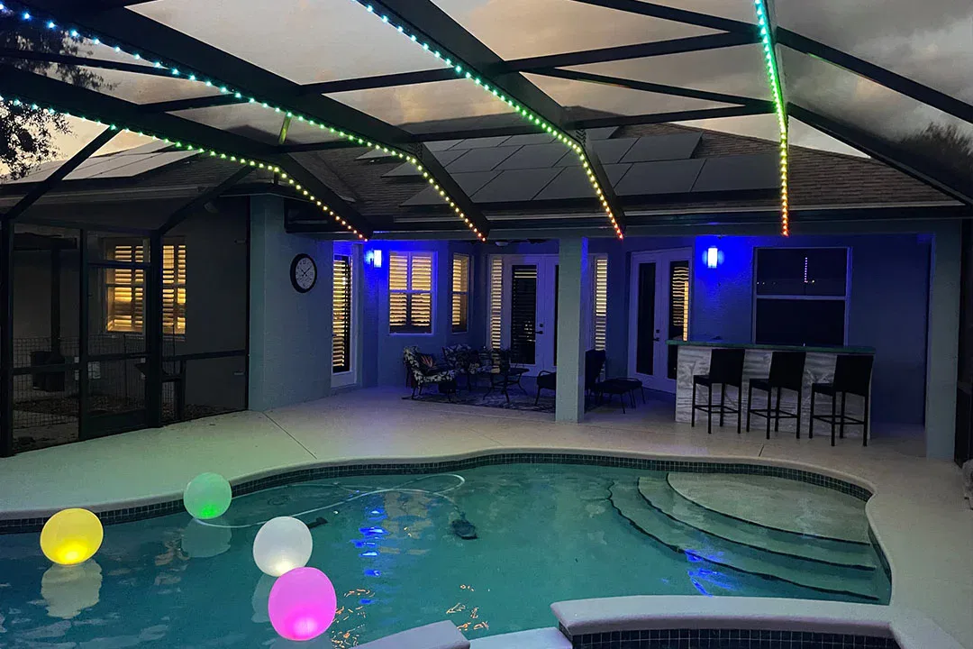 A backyard pool at dusk features floating, glowing balls in the water. A covered patio with colorful LED lights, bar stools, and lounge seating is visible in the background.