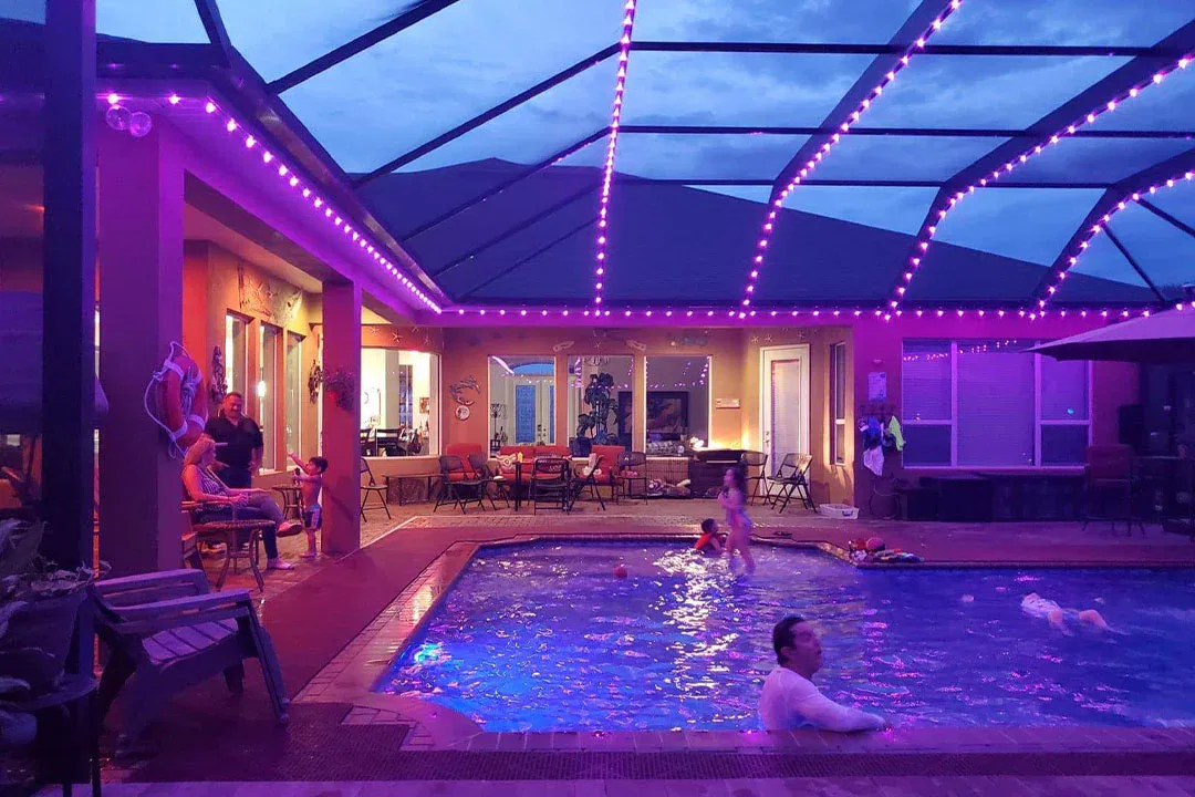People relax and swim in a backyard pool at dusk, surrounded by purple LED lights. Some are sitting by the poolside while others swim, and the enclosed patio area glows warmly through the windows.