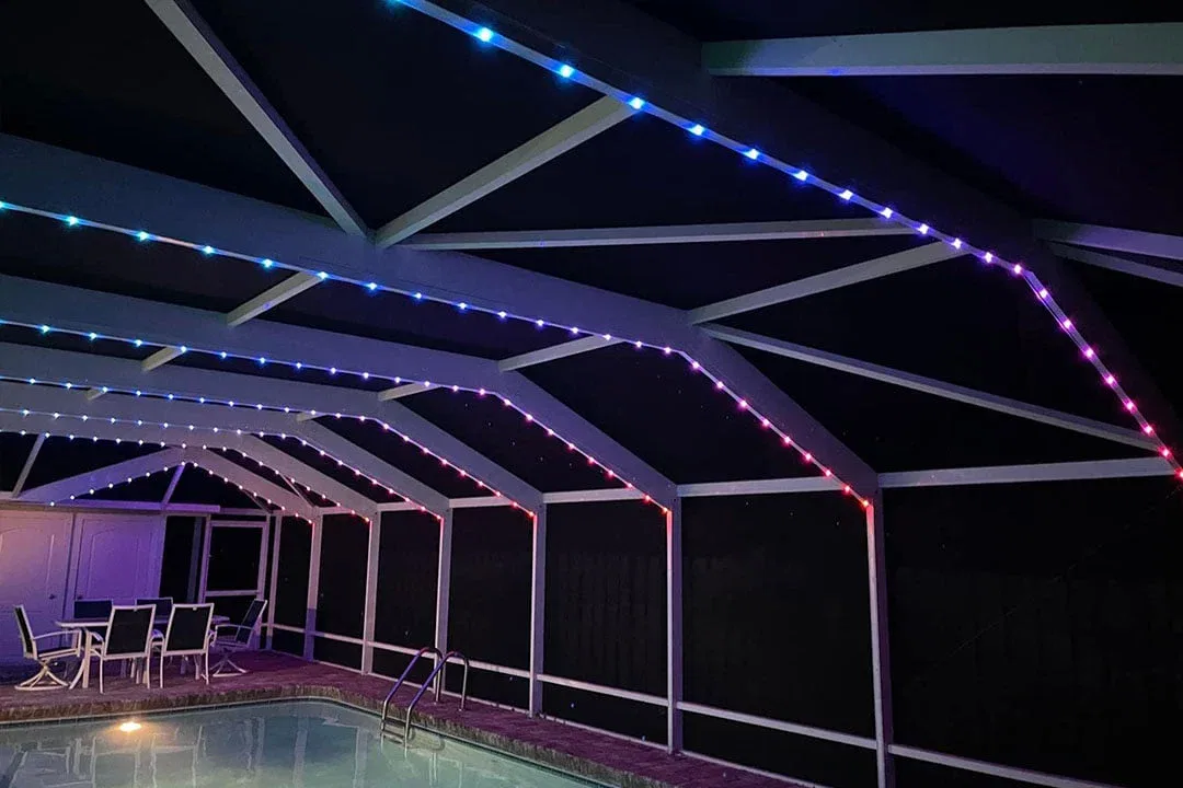 A screened-in patio with a pool features LED strip lights overhead glowing in blue, purple, and red. Patio furniture is arranged near the pool against the far wall.