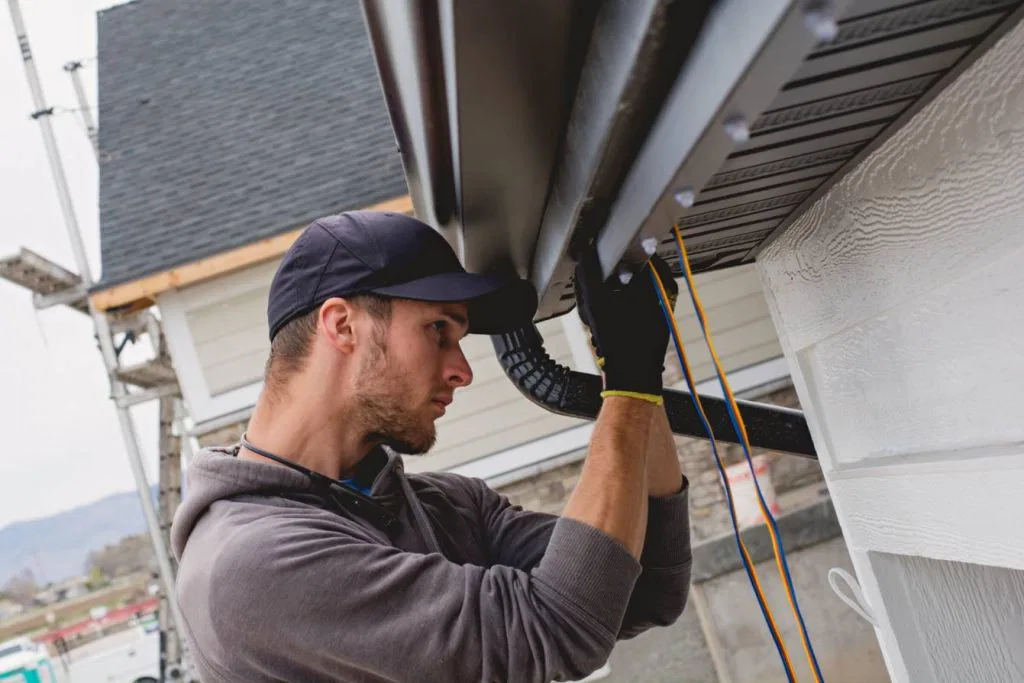 A man wearing a cap and gloves installs or repairs a rain gutter on the side of a house, using tools and cords, with a focus on the roofline and exterior siding.