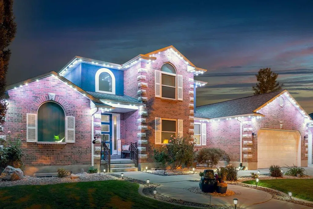 Two-story brick house with white trim, illuminated by bright white lights at dusk. The yard has manicured bushes, a curved walkway, and a lit driveway leading to a double garage. The sky is deep blue with pink clouds.