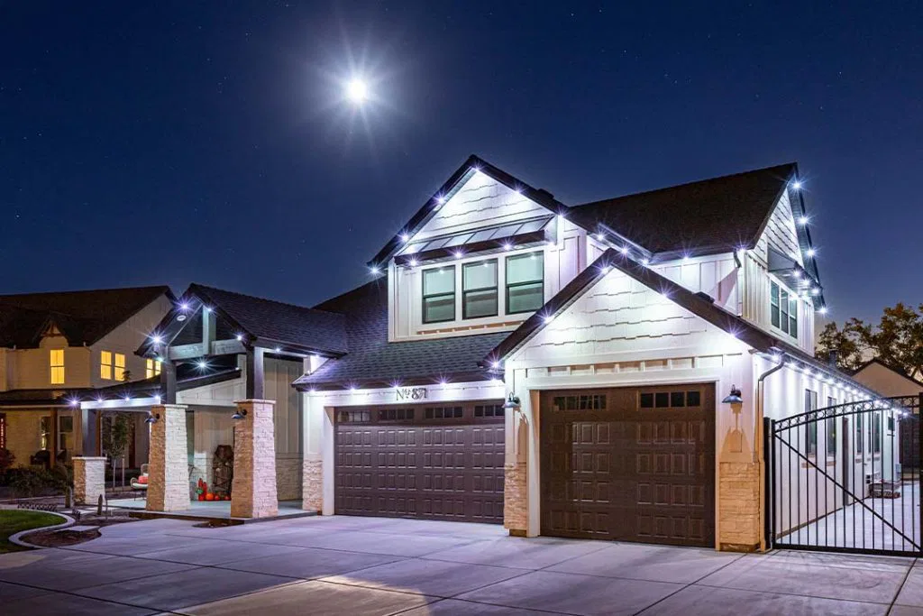 A modern two-story house at night, decorated with white string lights along the roofline. The driveway and garage doors are visible, and a bright full moon shines overhead in the clear sky.