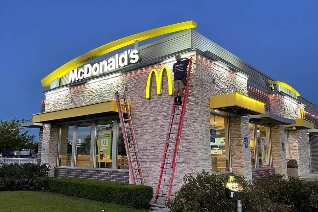 A McDonalds restaurant at dusk with two ladders propped against the building. A worker is on one ladder, adjusting lights above the entrance. The signature yellow arches and McDonalds sign are clearly visible.