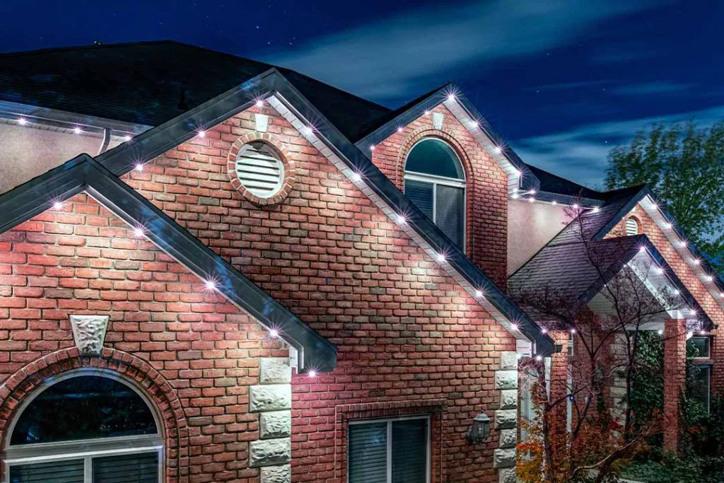 A brick house at night with white LED lights outlining its roof and gables, illuminating the exterior against a dark blue sky.