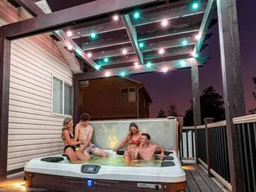 Four people in swimsuits relax in a hot tub on a deck at dusk, under a pergola decorated with string lights. Two women and two men are enjoying the water, with a house and trees visible in the background.