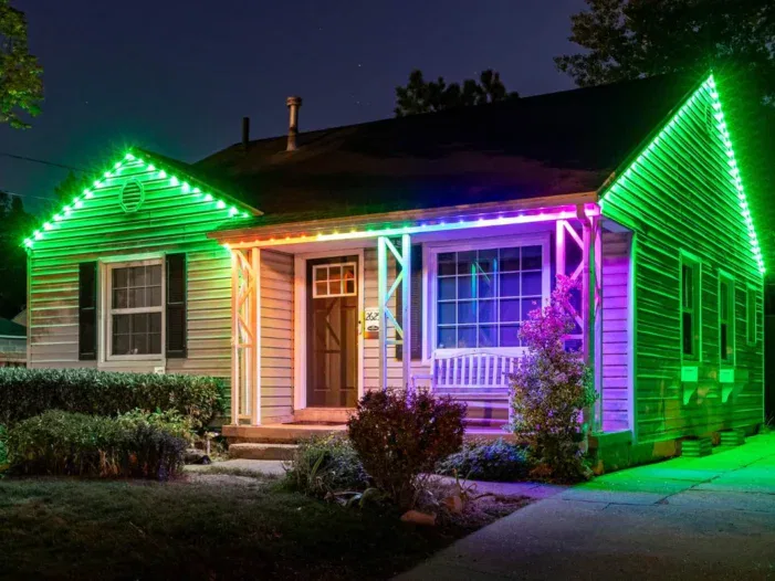 A single-story house at night is decorated with vibrant, multicolored LED lights along the roofline and porch pillars, creating a bright and festive appearance. Shrubs and plants surround the entrance.
