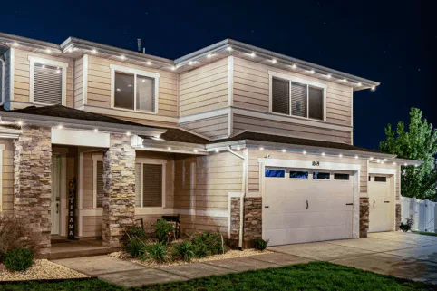 Two-story suburban house at night with stone accents, beige siding, a double garage, and warm outdoor lighting outlining the roof. Small shrubs and plants line the front yard.