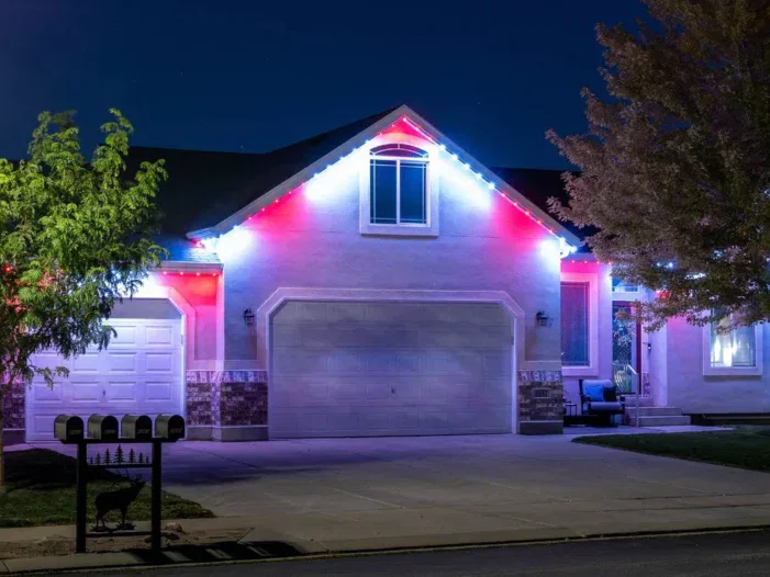 A suburban house at night with red, white, and blue lights outlining the roof. The driveway is empty, and trees frame the house on both sides. Three mailboxes are visible in the foreground.