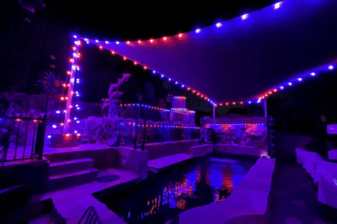 A backyard pool area at night, illuminated by red and blue string lights draped along a triangular canopy and fencing, reflecting off the water and creating a vibrant, colorful atmosphere.