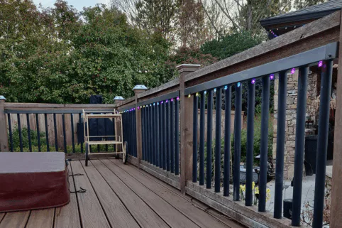A wooden deck with a railing lined with colorful string lights, a foldable chair, and a brown cushion. Trees and bushes fill the background, and a stone patio is visible beyond the railing.