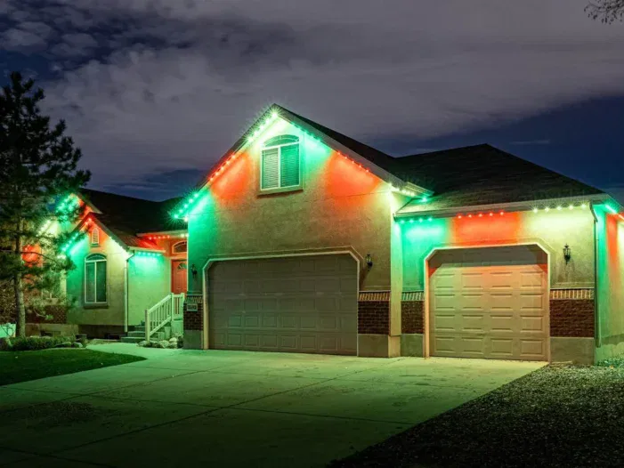 A house at night is decorated with red and green lights along the roofline, creating a festive holiday appearance. The driveway and yard are empty under a partly cloudy sky.
