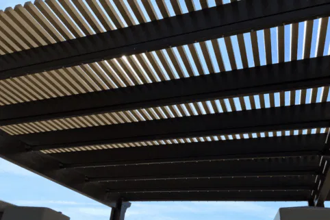 A close-up view from below of a pergola with evenly spaced, dark wooden slats casting striped shadows, with a blue sky and a few scattered clouds visible through the gaps.