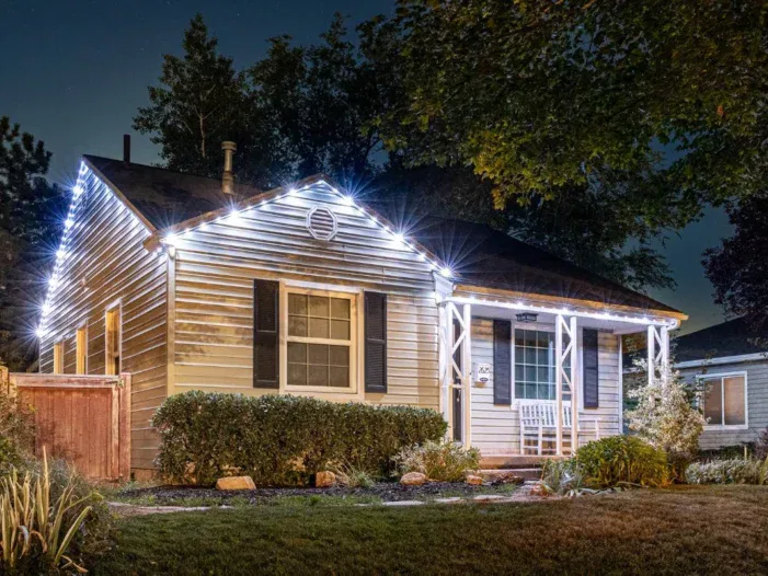 A cozy house at night with white holiday lights outlining the roof, bushes in front, a white bench on the porch, and a tree partially shading the yard.