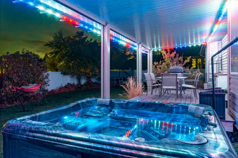 A hot tub with blue LED lights sits on a patio under a covered roof adorned with colorful string lights. Nearby are an outdoor dining table and chairs, with a hammock and trees visible in the backyard at night.
