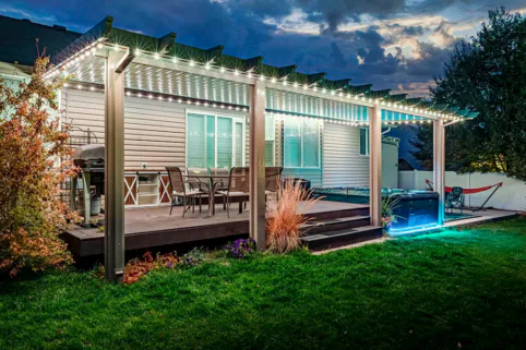 A backyard patio at dusk features a pergola with string lights, outdoor dining furniture, a hot tub with blue lighting, and a barbecue grill, surrounded by green grass and landscaping.