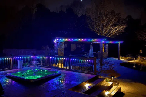 A backyard at night features a glowing hot tub on a wooden deck, with colorful LED lights outlining the deck rails and a nearby covered patio. Trees and landscaping are dimly visible in the background.