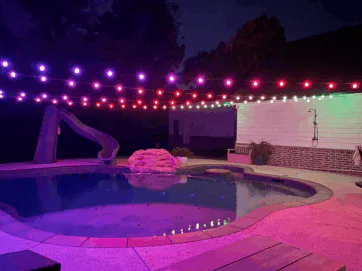 A backyard pool at night is lit by strings of purple, red, and white lights overhead. A slide leads into the pool, and a small rock waterfall feature sits beside it. The area is quiet and empty.