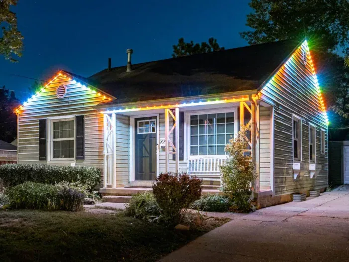 A small, single-story house at night with colorful LED lights outlining the roof, a front porch with a white bench, shrubs, and a paved walkway leading to the door.