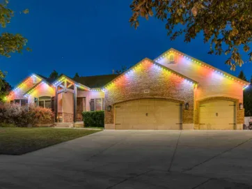 A large brick house with three beige garage doors is lit up at night by colorful string lights outlining the roof. The driveway and front yard are visible under the evening sky.