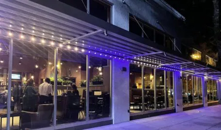 A modern restaurant exterior at night with large glass windows, warm interior lighting, visible diners, and a sidewalk illuminated by purple and white string lights under an awning.