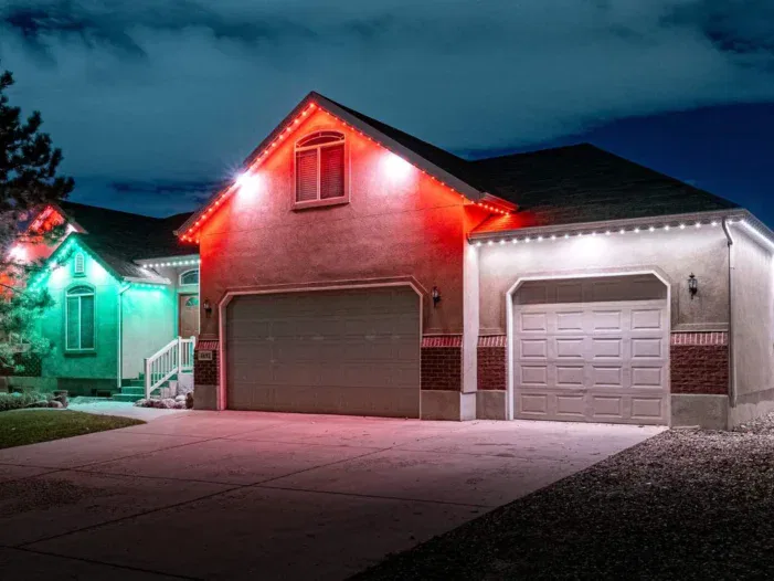 A house at night with garage doors, roof edges lit by red, white, and green LED lights, highlighting the outline of the building under a partly cloudy sky.