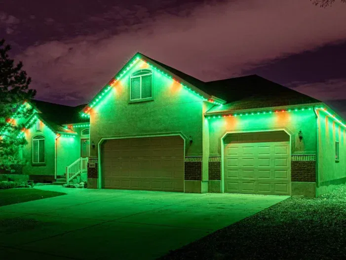 A suburban house at night with its roofline decorated in glowing green and red lights, illuminating the driveway and creating a festive atmosphere. The sky is partly cloudy.