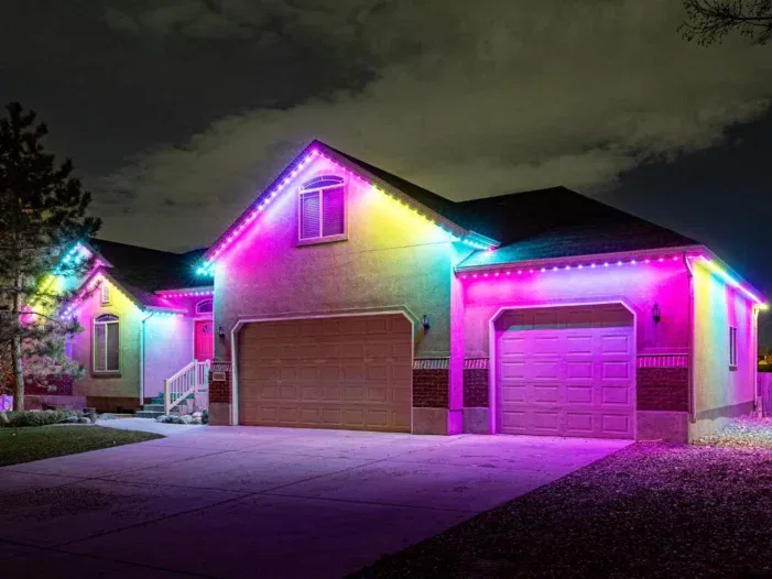 A house at night with colorful LED lights outlining the roof and garage, glowing in shades of purple, blue, pink, and yellow against a cloudy sky.