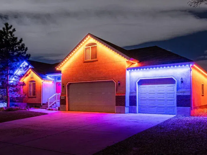 A single-story house at night with red and blue LED lights outlining the roof and garage, illuminating the driveway and creating a colorful glow against a cloudy sky.