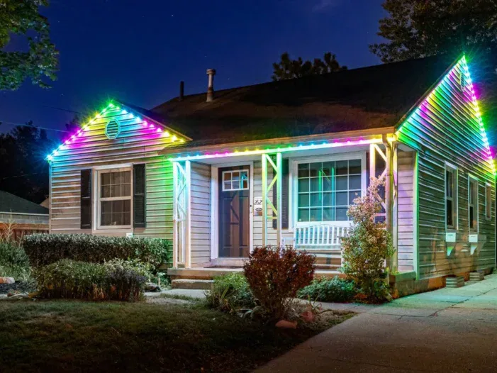 A single-story house at night with multicolored LED lights outlining the roof. The house has beige siding, dark shutters, a front porch with a white bench, and a small garden in front.
