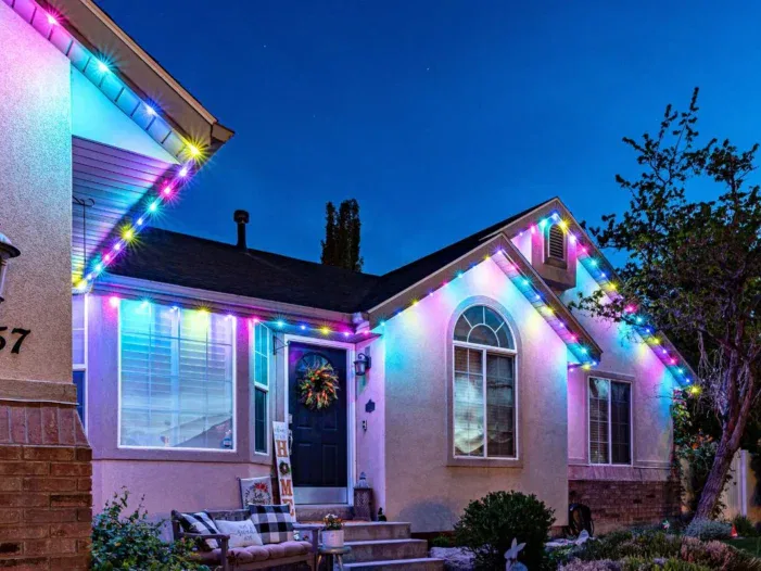 A house at dusk decorated with colorful lights along the roofline. The porch features a wreath on the door, a small bench, and a HOME sign. The sky is clear and trees are visible in the yard.