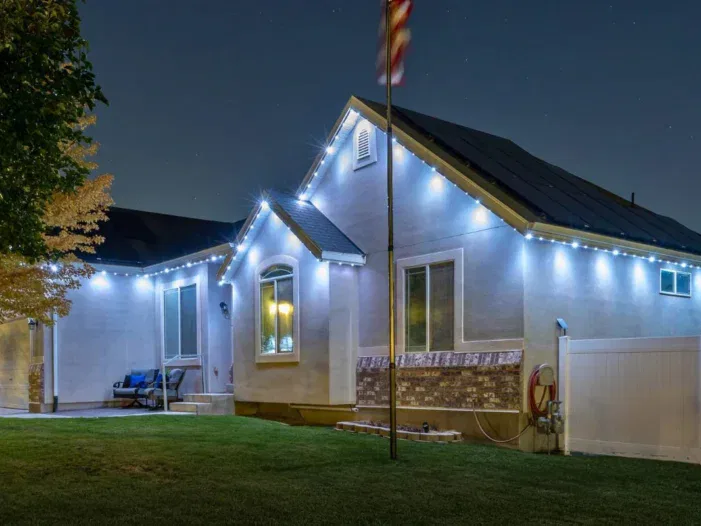 A single-story house at night with white exterior walls, decorative stone trim, and blue LED lights outlining the roof. The yard is neatly manicured, and a flagpole stands in front of the house.